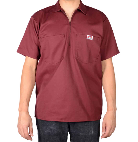 A person wearing a 1/2 Zipper Short Sleeve - Burgundy shirt with two chest pockets and a small logo patch, made in the USA, paired with dark jeans and standing against a white background.