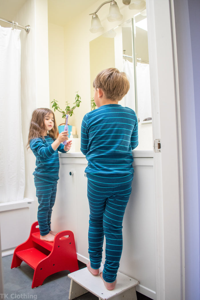 Two kids in matching blue striped pajamas by TK Clothing brushing teeth in a bright bathroom