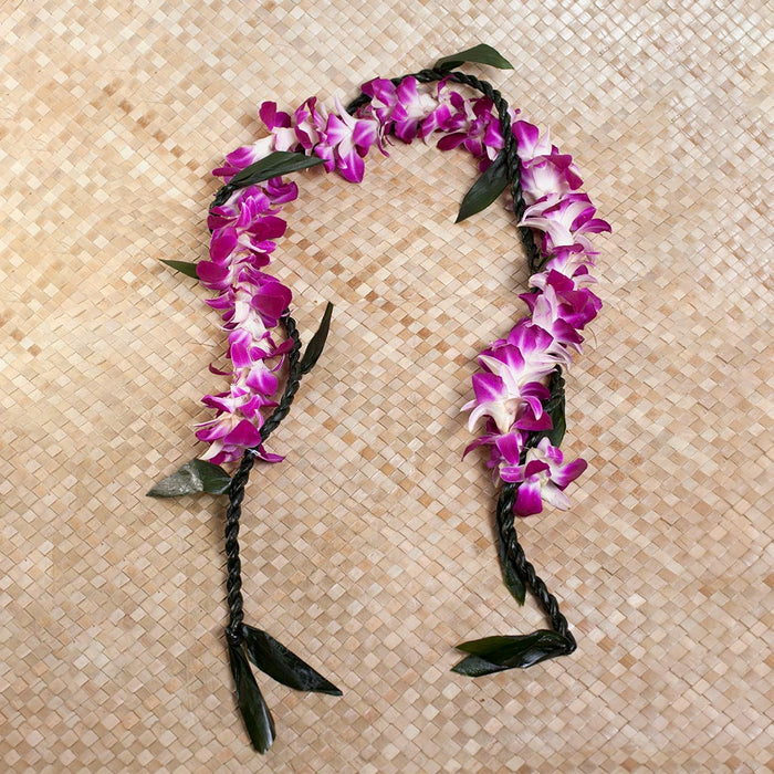 A traditional Hawaiian open-ended lei made of braided dark green ti leaves intertwined with vibrant purple dendrobium orchids, draped over a person's shoulders