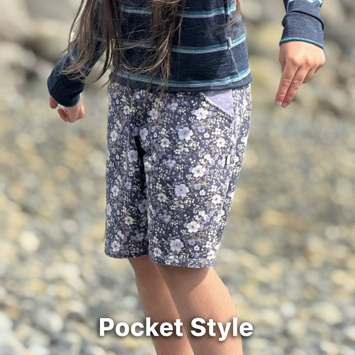 Child Wearing organic cotton purple flowery shorts on a pebble beach. Shorts made by TK Clothing