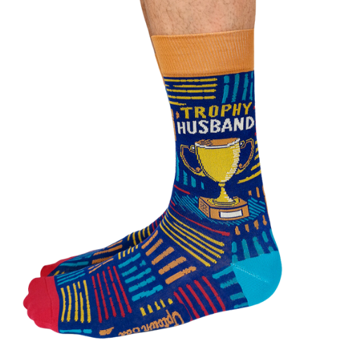 A pair of socks with a playful design featuring bold text that reads "Trophy Husband" in large, gold and white capital letters. The background is a deep, vibrant blue with small, scattered trophy icons in yellow and gold, adding a fun, humorous touch. The socks are mid-calf length and made of soft, stretchy material, perfect for a quirky gift or personal wear.