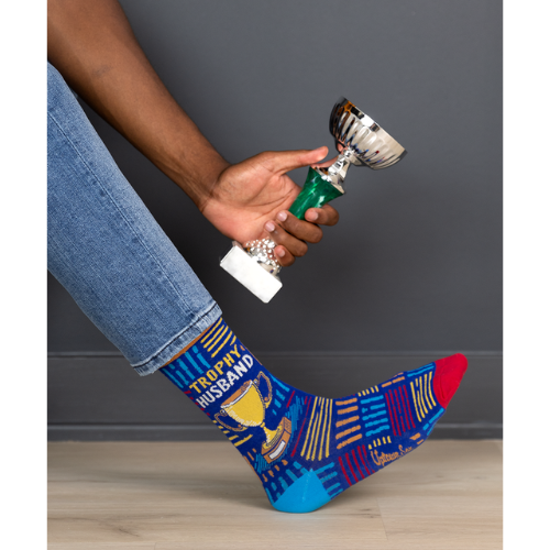 A pair of socks with a playful design featuring bold text that reads "Trophy Husband" in large, gold and white capital letters. The background is a deep, vibrant blue with small, scattered trophy icons in yellow and gold, adding a fun, humorous touch. The socks are mid-calf length and made of soft, stretchy material, perfect for a quirky gift or personal wear.