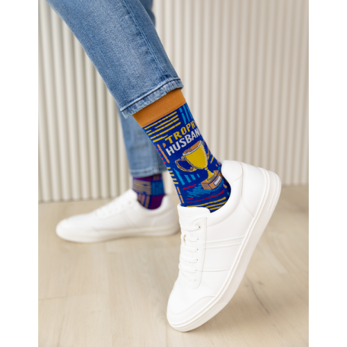 A pair of socks with a playful design featuring bold text that reads "Trophy Husband" in large, gold and white capital letters. The background is a deep, vibrant blue with small, scattered trophy icons in yellow and gold, adding a fun, humorous touch. The socks are mid-calf length and made of soft, stretchy material, perfect for a quirky gift or personal wear.